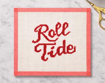 May include: A needlepoint canvas with the words "Roll Tide" stitched in red thread. The text is centered on a white background, framed by a coral-colored border. The canvas is rectangular.