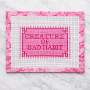 Op de afbeelding: Een roze borduurcanvas met de tekst "CREATURE OF BAD HABIT" in een bijpassende roze lijst. Het canvas is omgeven door een roze gemarmerde rand, geplaatst op een wit en grijs marmeren oppervlak.