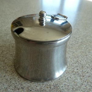 Puede incluir: Un tarro de condimento de metal plateado con tapa y un pequeño pico. El tarro tiene un acabado liso y brillante.