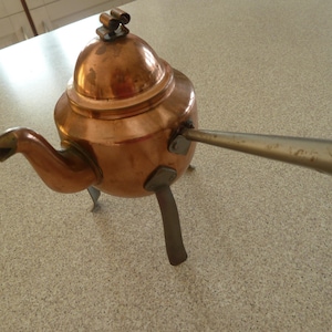 Könnte beinhalten: Eine Kupferteekanne mit einem gewölbten Deckel und einem langen Ausguss. Die Teekanne hat drei Metallbeine und einen langen Metallgriff.