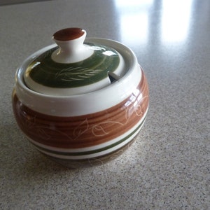 Puede incluir: Un azucarero de cerámica blanca con tapa. El tazón tiene rayas marrones y verdes y un diseño floral. La tapa tiene un pomo marrón y un diseño verde y blanco.