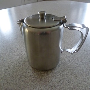 Może przedstawiać: Srebrny czajnik ze stali nierdzewnej z pokrywką na zawiasach i zakrzywionym uchwytem.