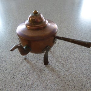 Puede incluir: Una tetera de cobre pequeña con tapa y asa. La tetera tiene tres patas y está sobre una superficie gris claro.