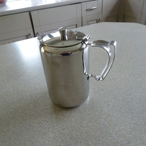 May include: A silver-colored, cylindrical pitcher with a lid and handle. The pitcher is made of a shiny metal and sits on a light-colored countertop. The handle has a decorative design.