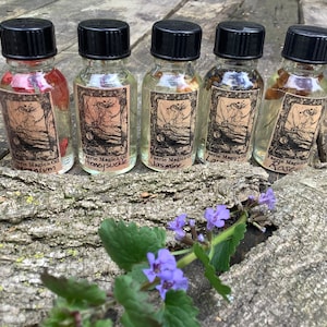 May include: Five small glass bottles with black caps, each filled with a clear liquid and containing dried flowers. The bottles are labeled "Faerie Magick Oil" with different flower names: "Geranium", "Honeysuckle", "Jasmine", "Lavender", and "Rose".