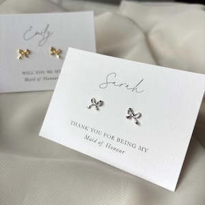 Puede incluir: Dos tarjetas blancas con el texto "Will you be my Maid of Honour" y "Thank you for being my Maid of Honour". Las tarjetas tienen los nombres "Emily" y "Sarah" escritos en cursiva. Cada tarjeta tiene un par de pendientes de lazo plateado.