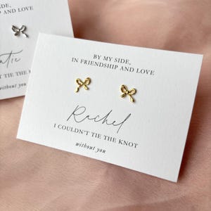 以下が含まれることがあります： ゴールドのボウタイのイヤリングと「By my side, in friendship and love」と「Rachel I couldn't tie the knot without you」のテキストが書かれた白いカード。
