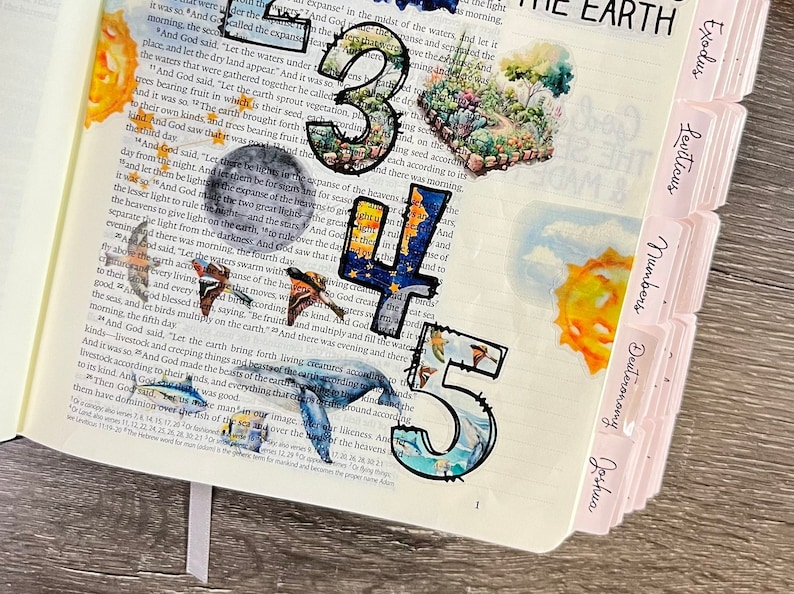 K&ouml;nnte beinhalten: Nahaufnahme einer Bibel mit bunten Papierschnitten der Zahlen 3, 4 und 5. Die Zahlen sind mit Bildern aus der Natur verziert, darunter eine Sonne, Wolken und ein Wal. Die Bibel hat rosa Laschen mit den Namen der B&uuml;cher der Bibel, darunter Exodus, Levitikus, Numeri, Deuteronomium und Josua.
