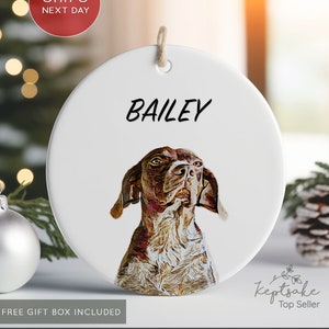 May include: A white, round ceramic ornament featuring a detailed portrait of a dog with brown and white fur. The name "BAILEY" is printed in black above the dog's image. A jute string is attached for hanging.
