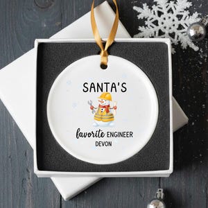 May include: White ceramic Christmas ornament with a gold ribbon, presented in a gift box. The ornament features a snowman dressed as an engineer, with the text "SANTA'S favorite ENGINEER DEVON".