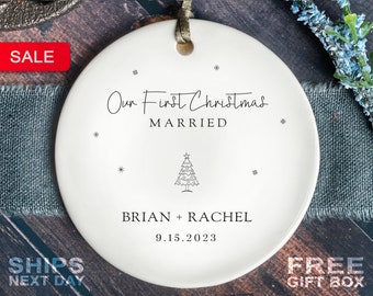 May include: A white ceramic Christmas ornament with the text "Our First Christmas MARRIED" and the names "BRIAN + RACHEL" along with the date "9.15.2023". The ornament has a small Christmas tree design and snowflake accents. A red "SALE" tag is visible.