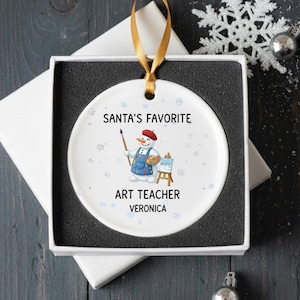 May include: A white ceramic Christmas ornament with a snowman artist design. The ornament reads "SANTA'S FAVORITE ART TEACHER VERONICA". It is presented in a white gift box with a gold ribbon for hanging.