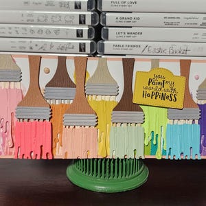 May include: A handmade greeting card featuring colorful paintbrushes with dripping paint in shades of pink, orange, yellow, green, blue, and purple. The card includes the text "You paint my world with HAPPINESS."