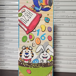 May include: A handmade greeting card featuring cartoon characters and cereal. The card includes a toucan, a tiger, and a rabbit, with the number 16. The cereal box reads "HB2U". Colorful cereal pieces and flowers are scattered across the card.