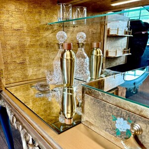 Puede incluir: Un mueble bar dorado con superficie de espejo que contiene dos cocteleras de lat&oacute;n con tapas de madera, dos decantadores de cristal transparente con patrones facetados, dos vasos de cristal y un colador de c&oacute;ctel de metal en un estante dorado.