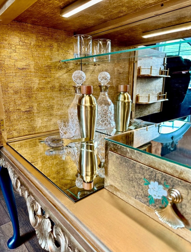 Puede incluir: Un mueble bar dorado con superficie de espejo que contiene dos cocteleras de lat&oacute;n con tapas de madera, dos decantadores de cristal transparente con patrones facetados, dos vasos de cristal y un colador de c&oacute;ctel de metal en un estante dorado.