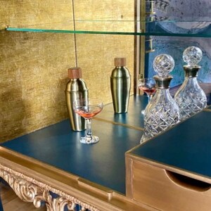 Puede incluir: Un mueble bar azul oscuro con adornos tallados en color dorado, que incluye dos cocteleras de metal dorado con tapas de madera, dos decantadores de cristal tallado transparente con tapones esf&eacute;ricos y dos copas con l&iacute;quido rosa.