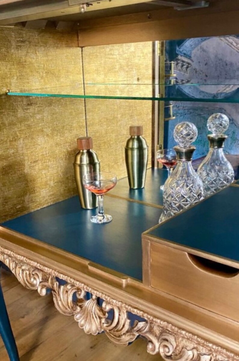 Puede incluir: Un mueble bar azul oscuro con adornos tallados en color dorado, que incluye dos cocteleras de metal dorado con tapas de madera, dos decantadores de cristal tallado transparente con tapones esf&eacute;ricos y dos copas con l&iacute;quido rosa.