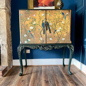 Puede incluir: Un armario rectangular de dos puertas con fondo dorado, pintado con p&aacute;jaros azules, flores blancas y ramas marrones. El mueble se apoya sobre cuatro patas cabriol&eacute; de color verde oscuro con detalles tallados y un motivo central de concha.