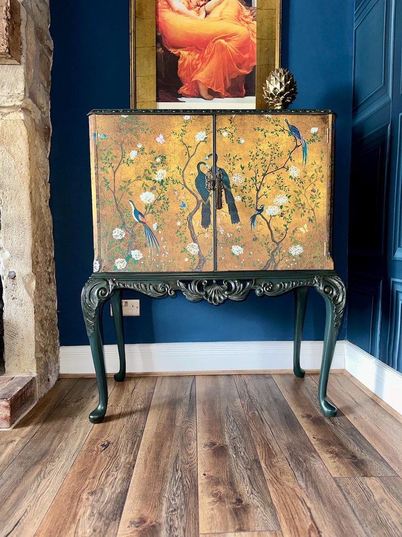 Puede incluir: Un armario rectangular de dos puertas con fondo dorado, pintado con p&aacute;jaros azules, flores blancas y ramas marrones. El mueble se apoya sobre cuatro patas cabriol&eacute; de color verde oscuro con detalles tallados y un motivo central de concha.