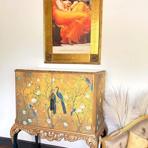 Puede incluir: Un armario rectangular de dos puertas con fondo dorado, p&aacute;jaros azules y ramas con flores blancas. El mueble tiene patas doradas talladas y una base oscura. Encima cuelga una l&aacute;mina enmarcada de una persona con un vestido naranja.