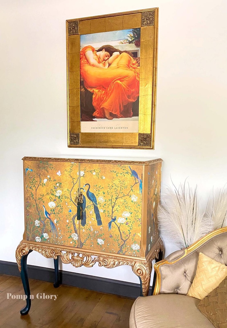 Puede incluir: Un armario rectangular de dos puertas con fondo dorado, p&aacute;jaros azules y ramas con flores blancas. El mueble tiene patas doradas talladas y una base oscura. Encima cuelga una l&aacute;mina enmarcada de una persona con un vestido naranja.