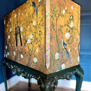 Puede incluir: Un armario alto con acabado dorado y una escena pintada de p&aacute;jaros, mariposas y ramas florecientes. La secci&oacute;n superior rectangular descansa sobre patas talladas de color verde oscuro con detalles curvos y bisagras visibles.