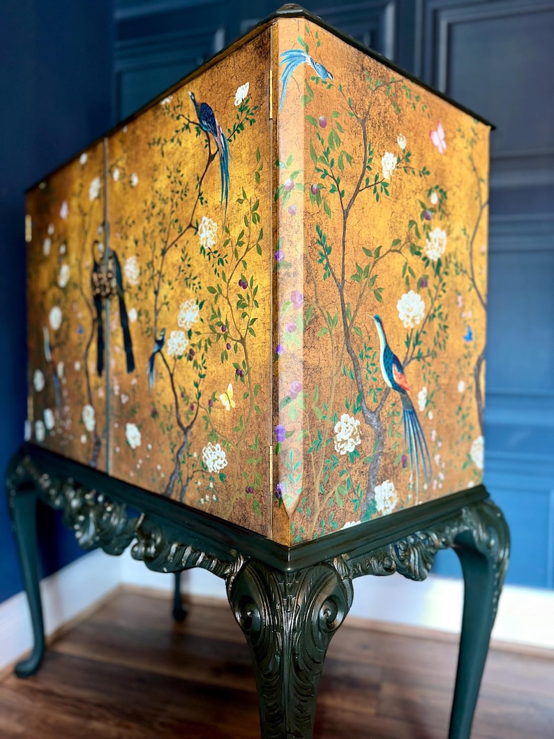Puede incluir: Un armario alto con acabado dorado y una escena pintada de p&aacute;jaros, mariposas y ramas florecientes. La secci&oacute;n superior rectangular descansa sobre patas talladas de color verde oscuro con detalles curvos y bisagras visibles.