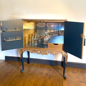 Puede incluir: Un mueble bar azul marino y dorado con dos puertas abiertas, que cuenta con un panel trasero de espejo, un estante de cristal y almacenamiento interior dorado. El exterior tiene tallas doradas ornamentadas en el fald&oacute;n y las patas, con dos estantes.