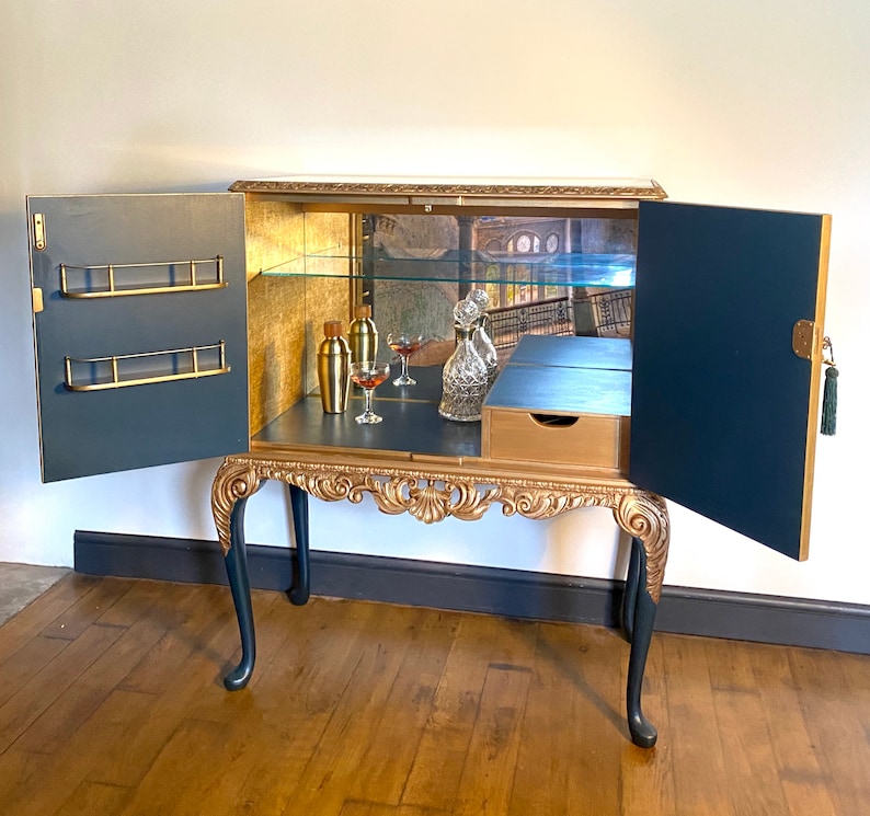 Puede incluir: Un mueble bar azul marino y dorado con dos puertas abiertas, que cuenta con un panel trasero de espejo, un estante de cristal y almacenamiento interior dorado. El exterior tiene tallas doradas ornamentadas en el fald&oacute;n y las patas, con dos estantes.