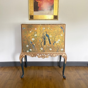 Puede incluir: Un gabinete rectangular con dos puertas sobre un fondo dorado, pintado con p&aacute;jaros azules, flores blancas y ramas oscuras. El mueble tiene molduras doradas ornamentadas y cuatro patas cabriol&eacute; de color gris oscuro con detalles dorados.