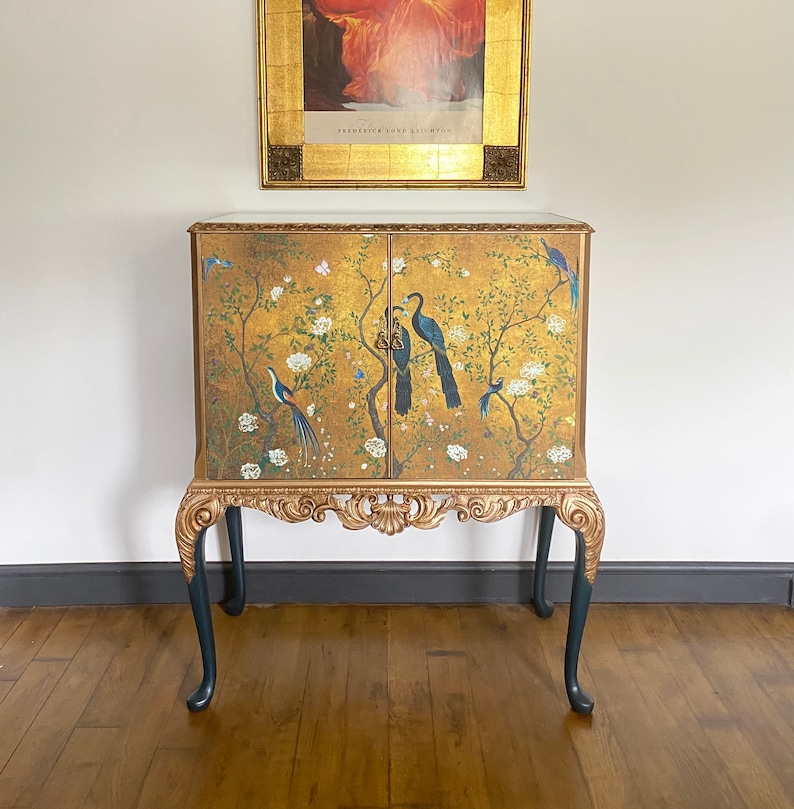 Puede incluir: Un gabinete rectangular con dos puertas sobre un fondo dorado, pintado con p&aacute;jaros azules, flores blancas y ramas oscuras. El mueble tiene molduras doradas ornamentadas y cuatro patas cabriol&eacute; de color gris oscuro con detalles dorados.
