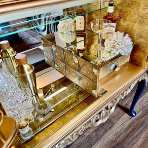 Puede incluir: Un mueble bar dorado con espejo, patas talladas, estantes de cristal y un caj&oacute;n con detalles florales pintados. Contiene cocteleras doradas, cristaler&iacute;a, una botella de Jack Daniel's Honey y adornos florales de color blanco.