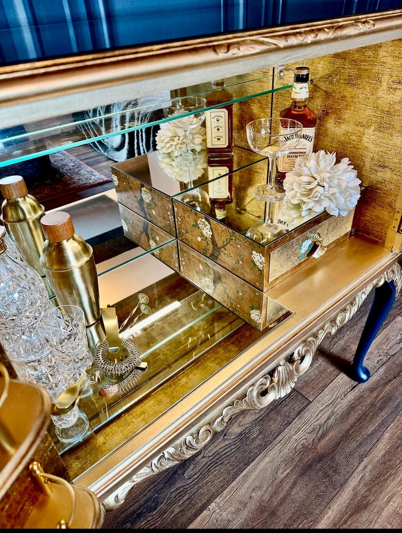 Puede incluir: Un mueble bar dorado con espejo, patas talladas, estantes de cristal y un caj&oacute;n con detalles florales pintados. Contiene cocteleras doradas, cristaler&iacute;a, una botella de Jack Daniel's Honey y adornos florales de color blanco.