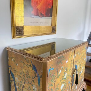 Puede incluir: Un armario dorado con tapa de espejo y dos puertas con un estampado de estilo chinoiserie de p&aacute;jaros azules, flores blancas y ramas. El mueble presenta bordes tallados ornamentados y una borla verde oscuro colgando de una llave met&aacute;lica.