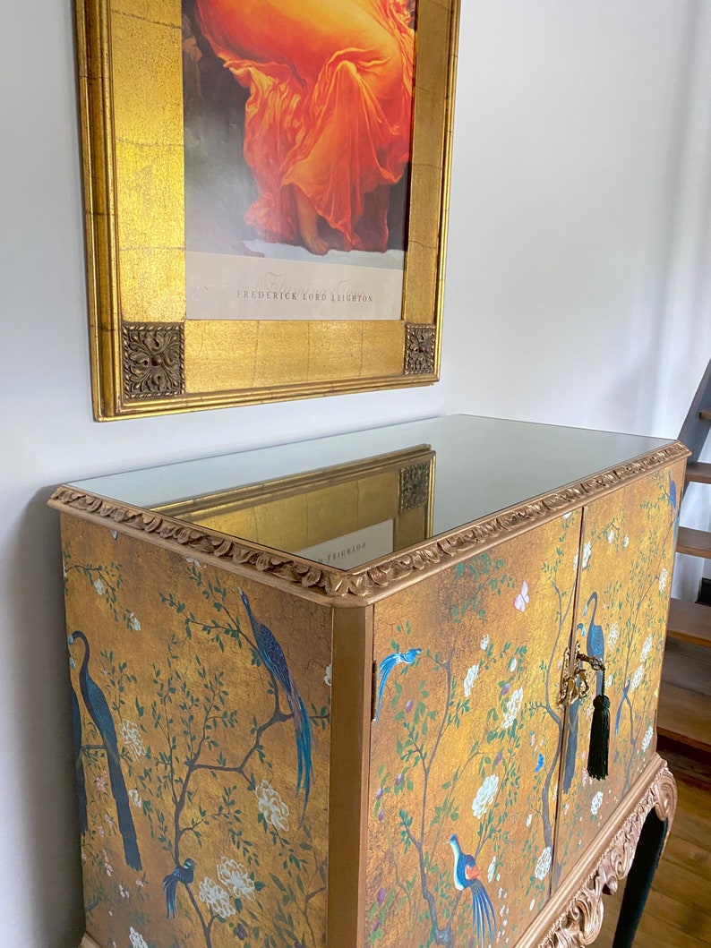 Puede incluir: Un armario dorado con tapa de espejo y dos puertas con un estampado de estilo chinoiserie de p&aacute;jaros azules, flores blancas y ramas. El mueble presenta bordes tallados ornamentados y una borla verde oscuro colgando de una llave met&aacute;lica.