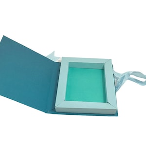 Op de afbeelding: Een lichtblauwe geschenkdoos met een wit frame en een lichtblauw lint als handvat. De doos is open en de binnenkant is bekleed met lichtgroen papier.