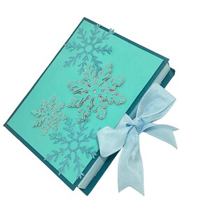 Op de afbeelding: Een tealblauwe cadeaudoos met een lichtblauw lint. De doos is versierd met zilveren glitter sneeuwvlokken.