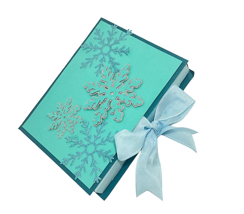 Op de afbeelding: Een tealblauwe cadeaudoos met een lichtblauw lint. De doos is versierd met zilveren glitter sneeuwvlokken.