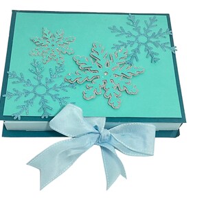 Op de afbeelding: Een turquoise blauwe geschenkdoos met een lichtblauw lint. De doos is versierd met zilveren glitter sneeuwvlokken.