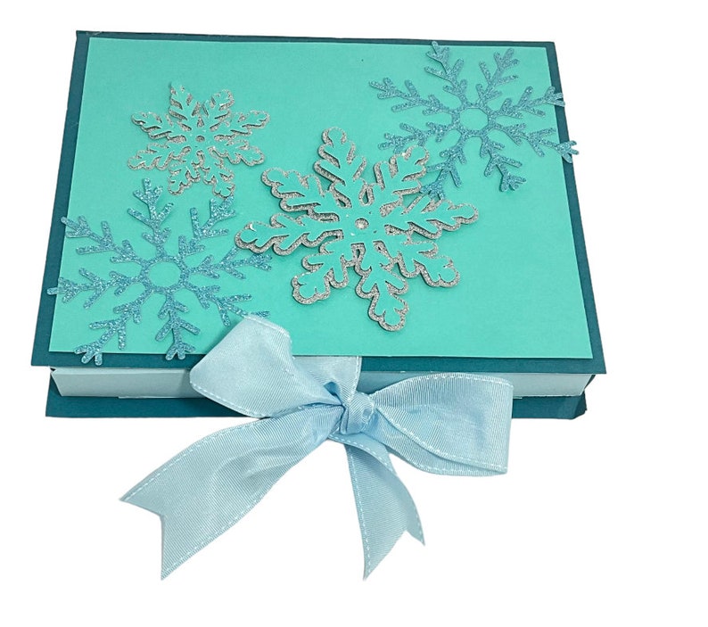 Op de afbeelding: Een turquoise blauwe geschenkdoos met een lichtblauw lint. De doos is versierd met zilveren glitter sneeuwvlokken.