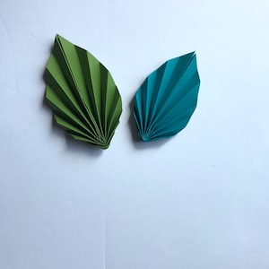Pode incluir: Duas folhas de origami, uma verde e outra azul, dobradas em forma de leque, sobre um fundo branco.