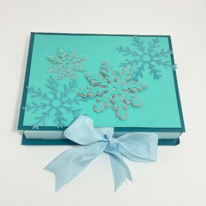 Op de afbeelding: Een turquoise blauwe cadeaudoos met een blauwe strik. De doos is versierd met zilveren glitter sneeuwvlokken.