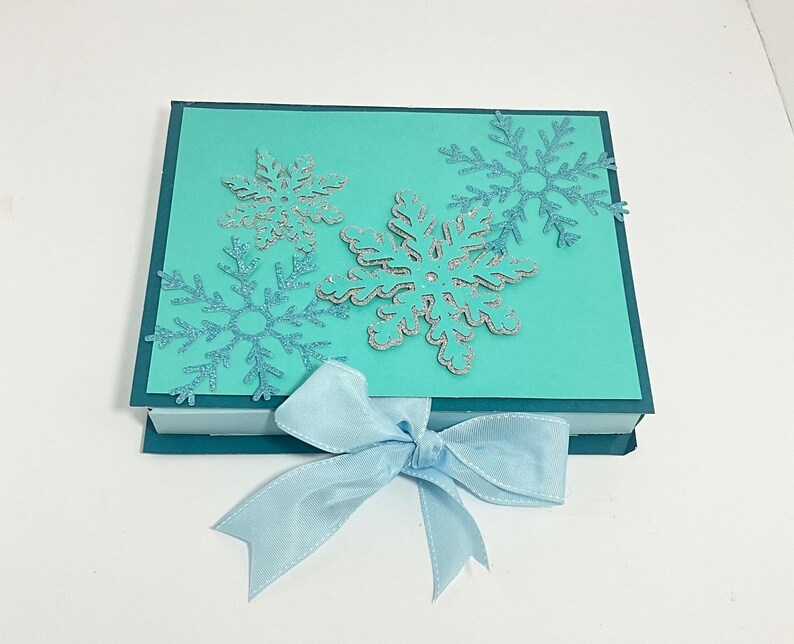 Op de afbeelding: Een turquoise blauwe cadeaudoos met een blauwe strik. De doos is versierd met zilveren glitter sneeuwvlokken.