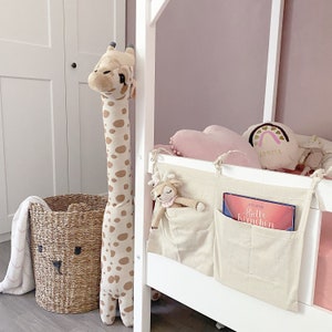 Könnte beinhalten: Ein weißes Hochbett mit einer rosa Wand dahinter. Das Bett hat einen weißen Stoff-Aufbewahrungsorganizer mit zwei Taschen. Der Organizer hat ein Buch in einer Tasche und ein Stofftier in der anderen. An der Bettstütze lehnt eine Stoffgiraffe.