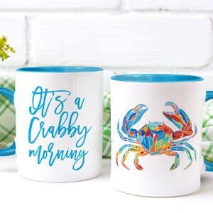 May include: Two white ceramic mugs with blue rims. One mug has the text "It's a Crabby Morning" in blue lettering. The other mug has a colorful illustration of a crab.