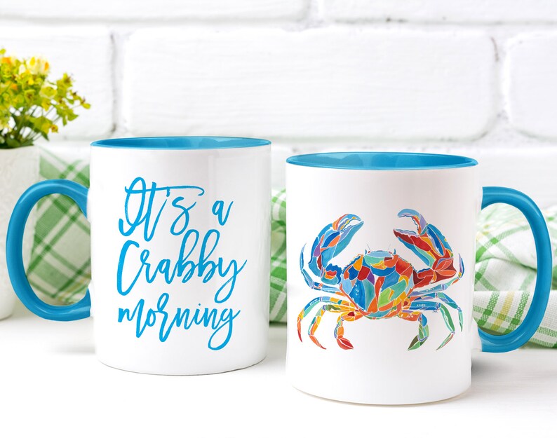 May include: Two white ceramic mugs with blue rims. One mug has the text "It's a Crabby Morning" in blue lettering. The other mug has a colorful illustration of a crab.