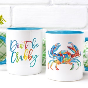 May include: Two white ceramic mugs with blue rims. One mug has the text "Don't be Crabby" in rainbow colors. The other mug has a colorful illustration of a crab.