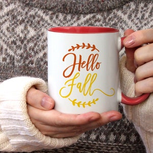 K&ouml;nnte beinhalten: Wei&szlig;e Keramiktasse mit rotem Rand und Henkel. Die Tasse hat ein Design mit der Aufschrift "Hello Fall" in orangefarbenen und gelben Buchstaben mit einem Blattdesign.