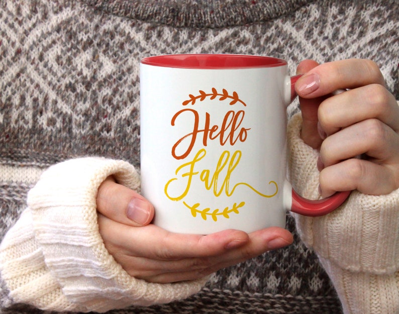 K&ouml;nnte beinhalten: Wei&szlig;e Keramiktasse mit rotem Rand und Henkel. Die Tasse hat ein Design mit der Aufschrift "Hello Fall" in orangefarbenen und gelben Buchstaben mit einem Blattdesign.